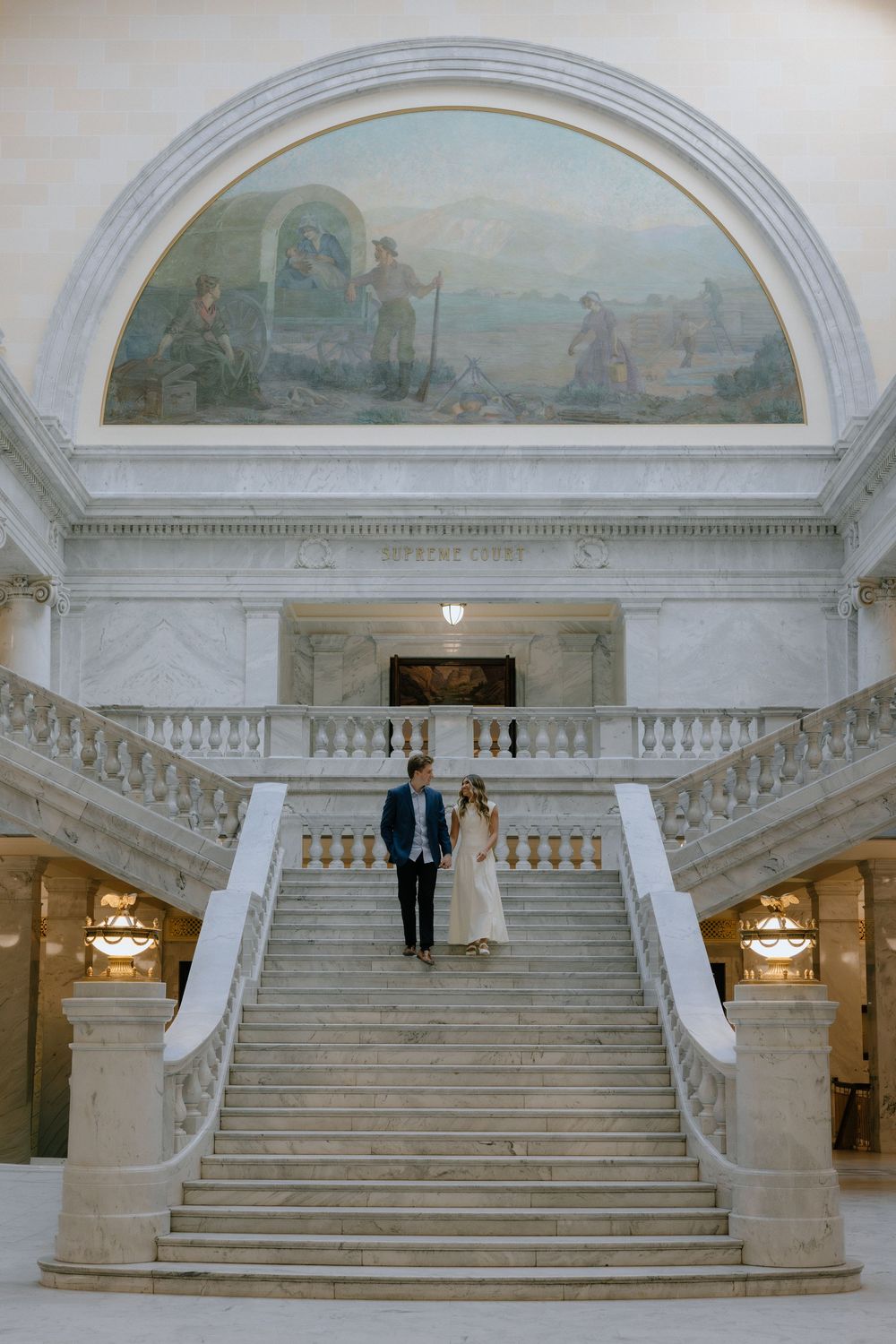 Couple poses on grand marble staircase beneath a classical mural in an elegant government building.