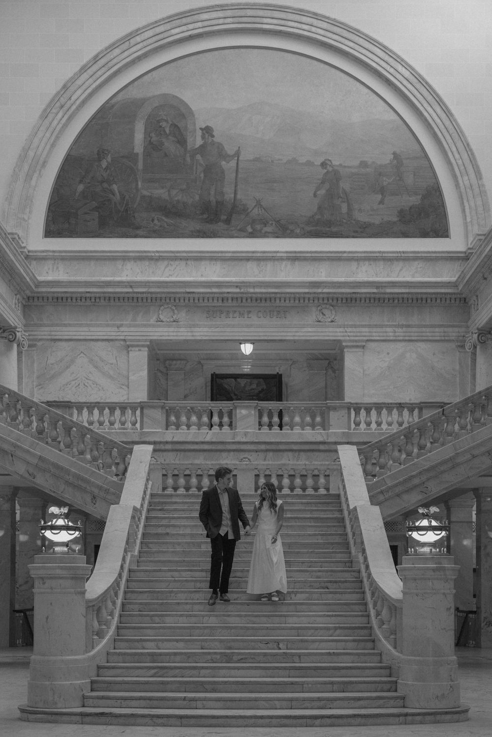 Black and white photo of couple holding hands on grand marble staircase beneath historic pioneer mural.