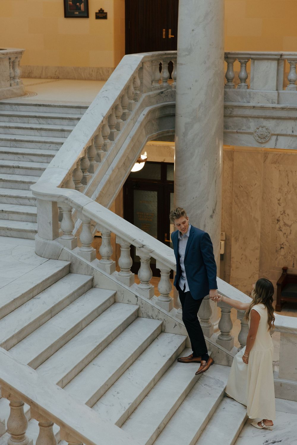 Couple holds hands on ornate marble staircase inside a historic neoclassical building.