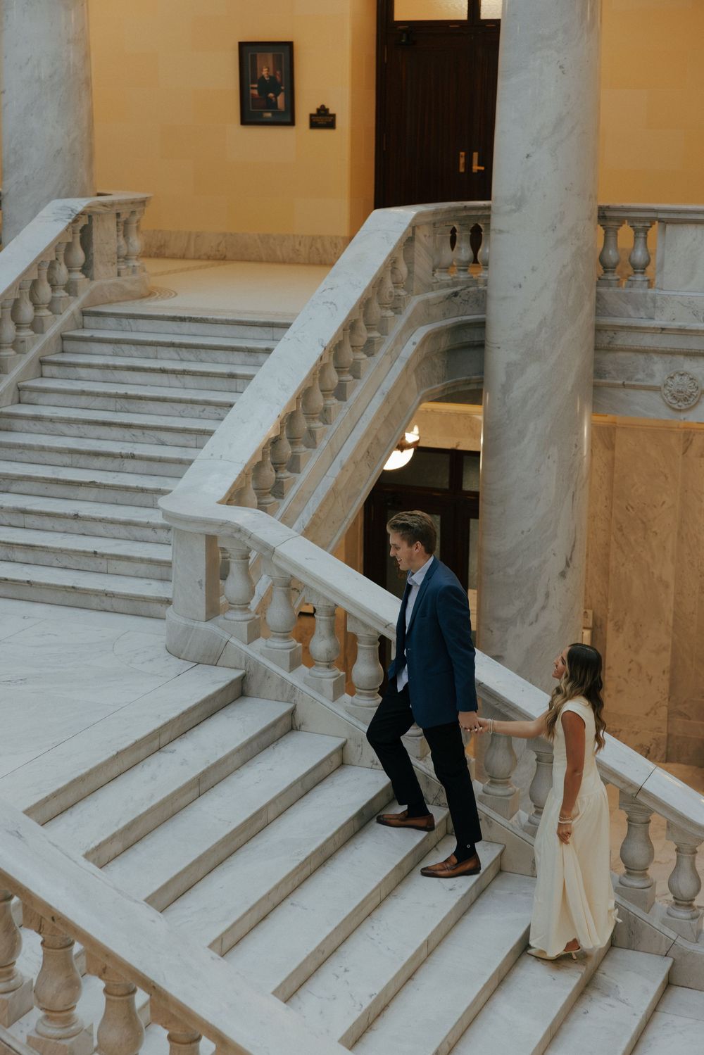 Couple holding hands while walking up elegant marble staircase inside a grand building.