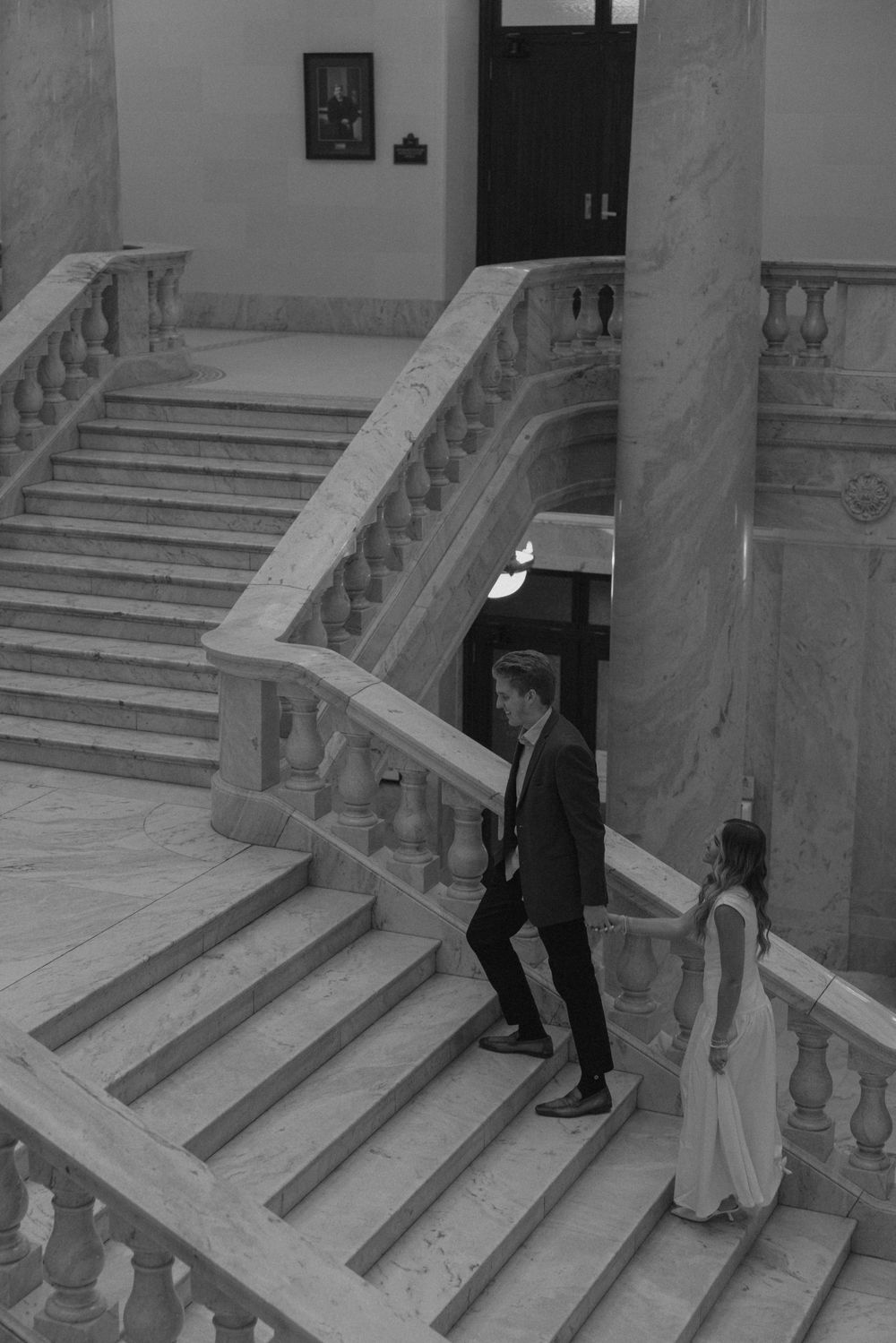 Black and white photo of couple walking down ornate marble staircase in historic building.