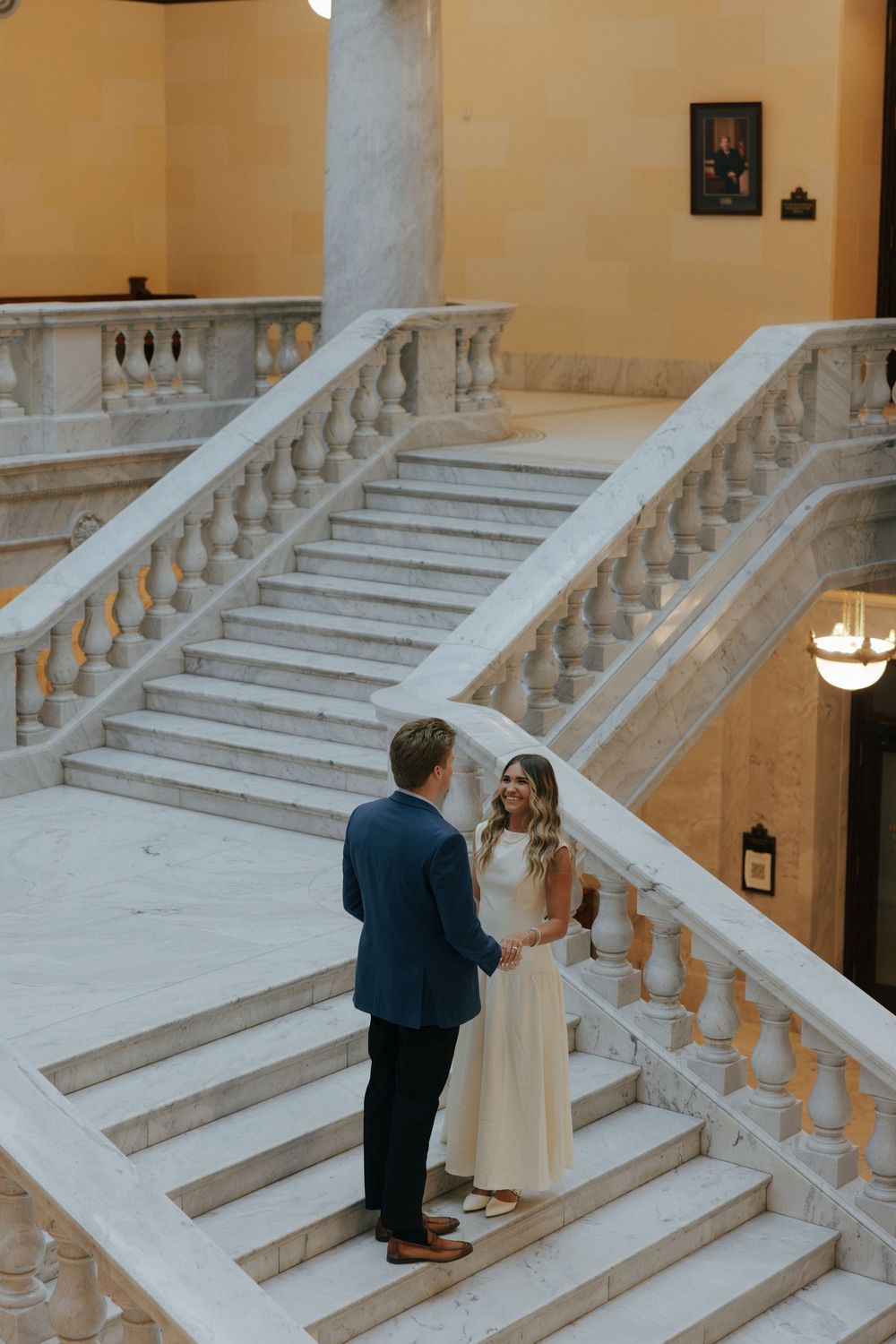 Couple holds hands on grand marble staircase inside elegant building during engagement session.