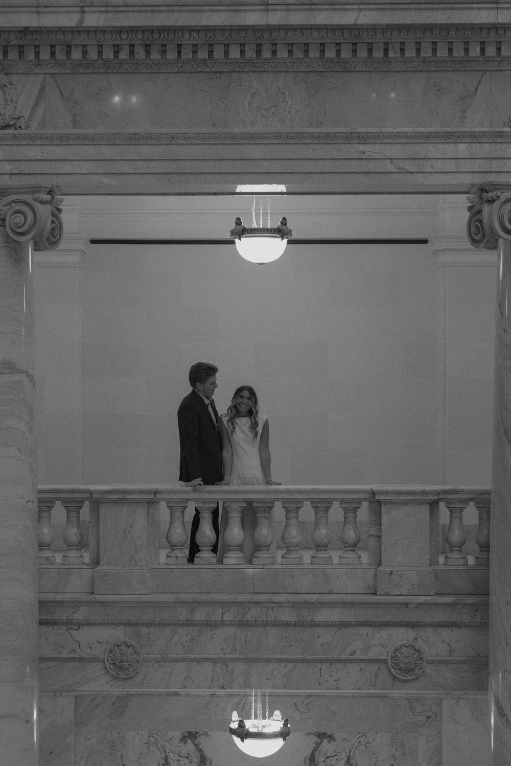 Black and white photo of a wedding couple standing on a grand marble balcony in an elegant building.