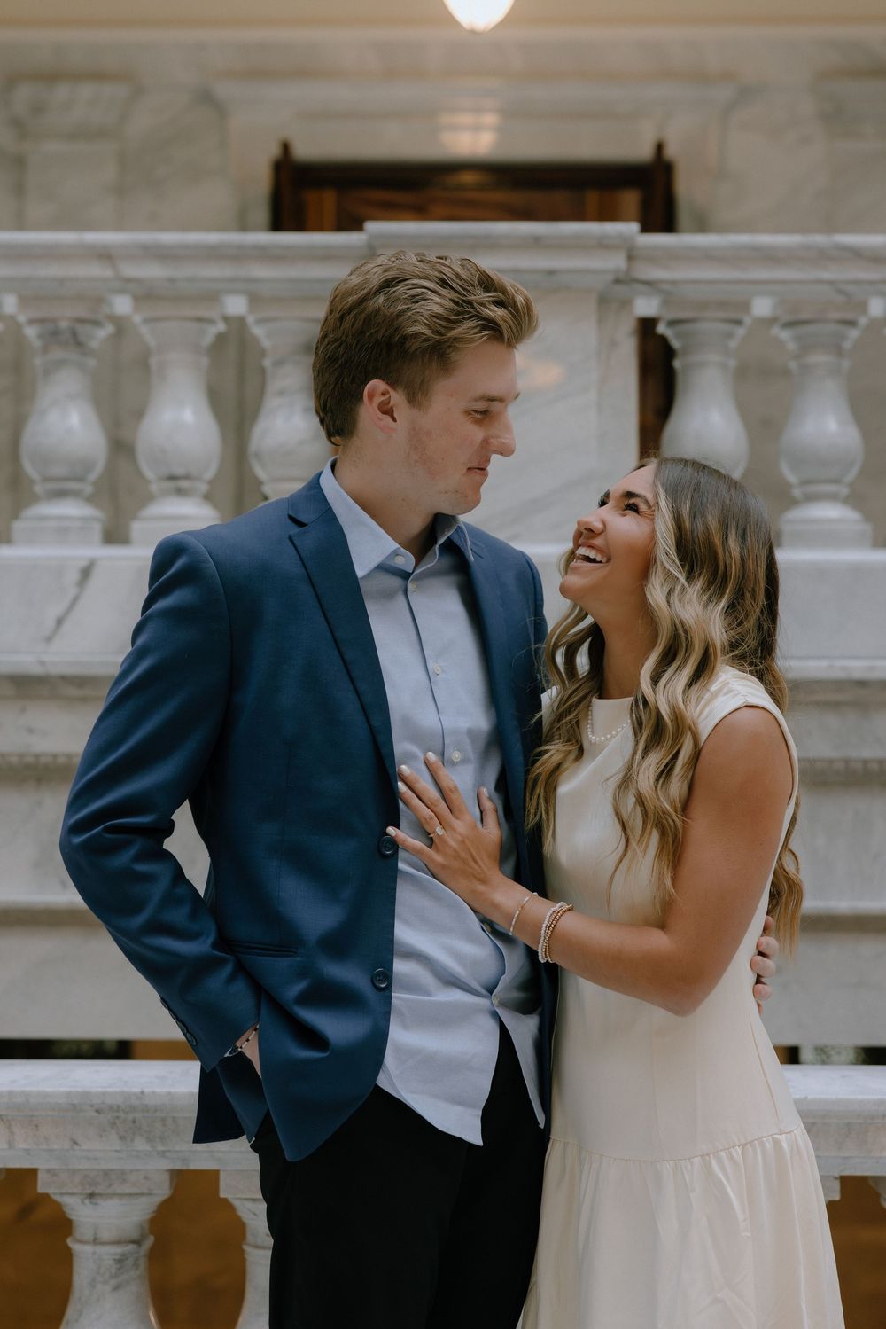 Couple laughing together in elegant marble building, man in navy suit, woman in white dress.