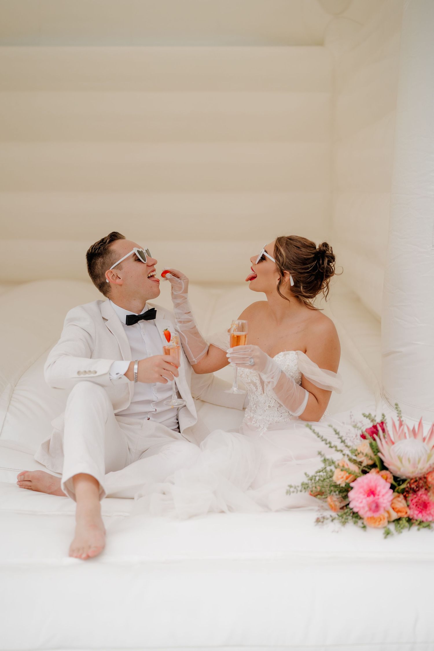 Bride and groom sitting in white bounce house, wearing fun glasses, holding champagne, with colorful floral bouquet nearby.