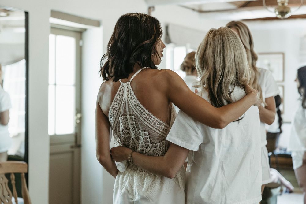 Bride in lace halter wedding dress embracing bridesmaids in a bright getting-ready room.