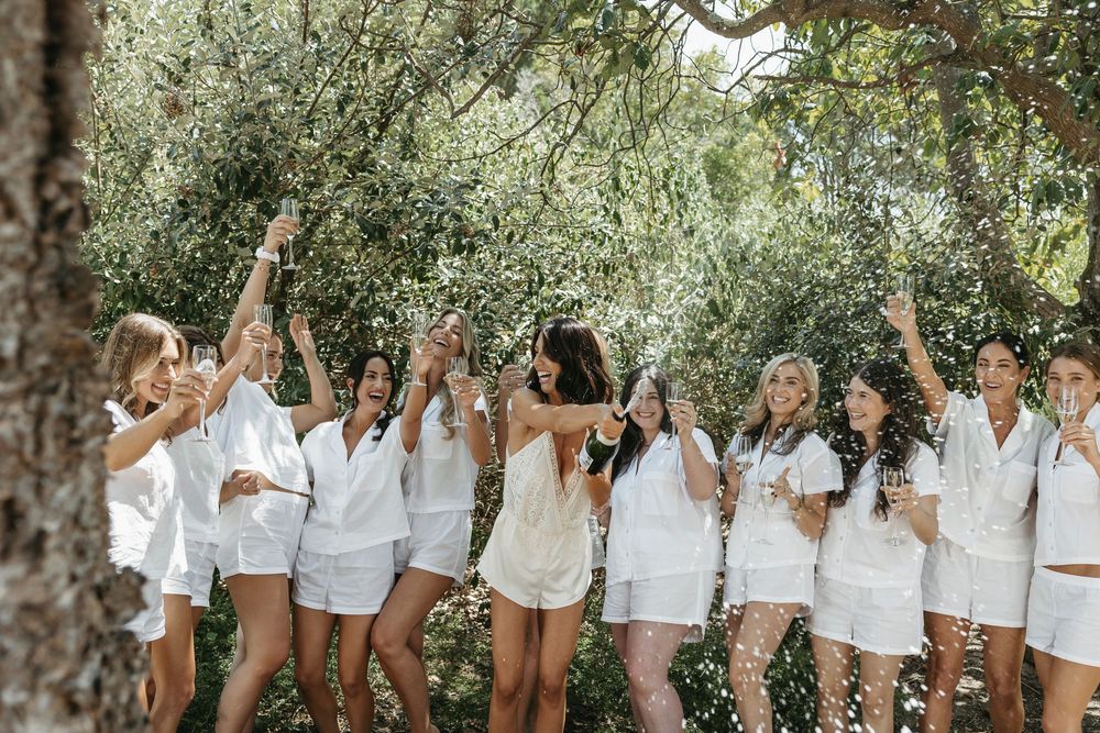 Bridal party in matching white outfits celebrating with champagne and confetti outdoors.