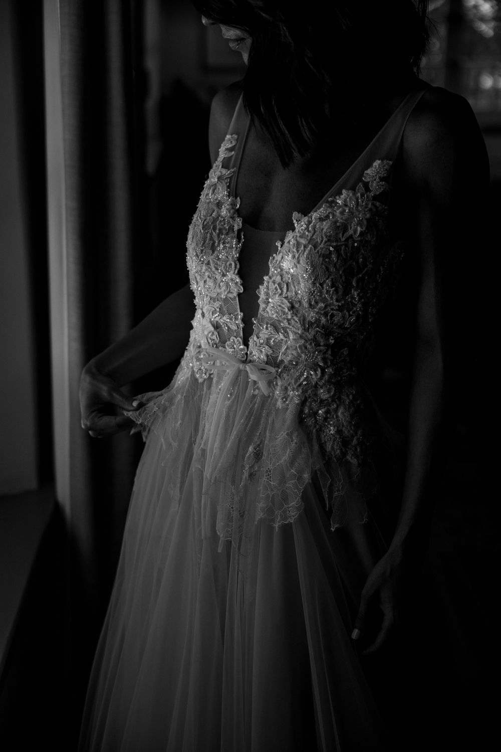 Black and white close-up of a bride in a floral lace bodice tulle wedding gown by a window.