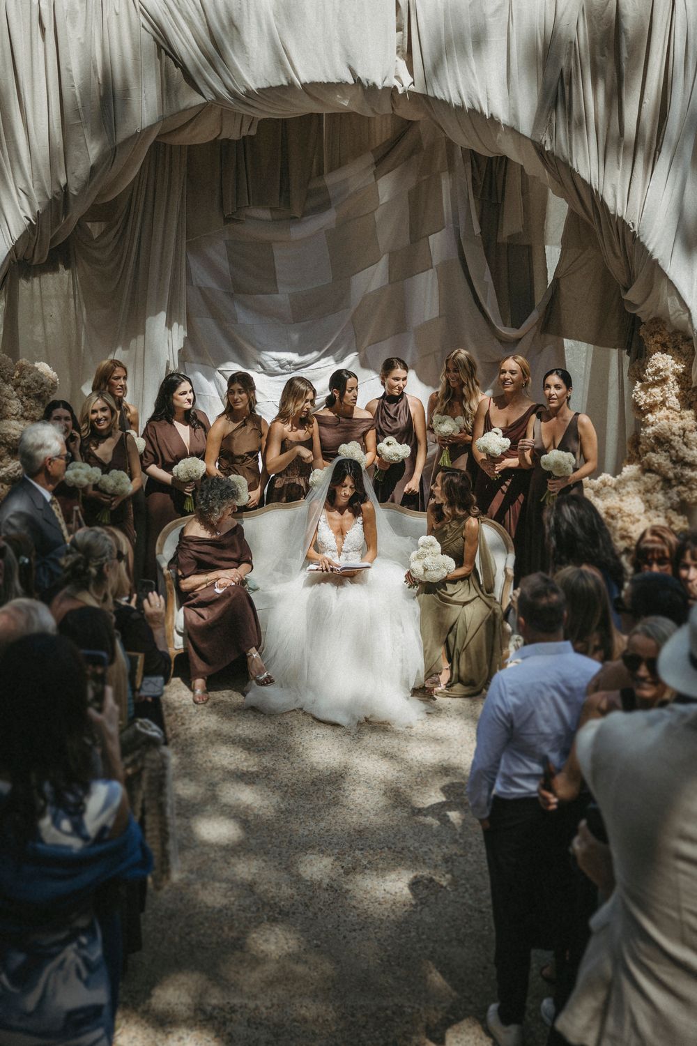 Bride in white gown reads vows seated under rustic fabric arch surrounded by bridesmaids.