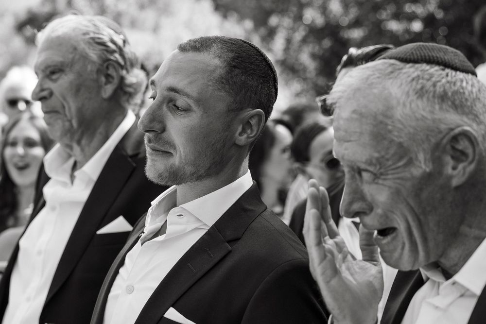 Emotional groom flanked by family members during outdoor wedding ceremony in black and white.