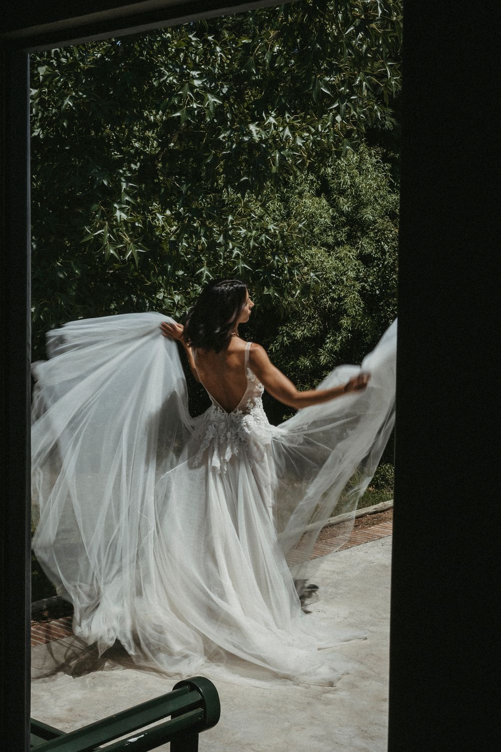 Bride in a flowing lace tulle wedding dress twirling outdoors surrounded by lush greenery.