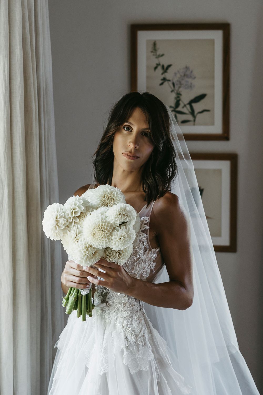 Bride holding a white dahlia bouquet wearing a lace wedding gown and veil by a window.