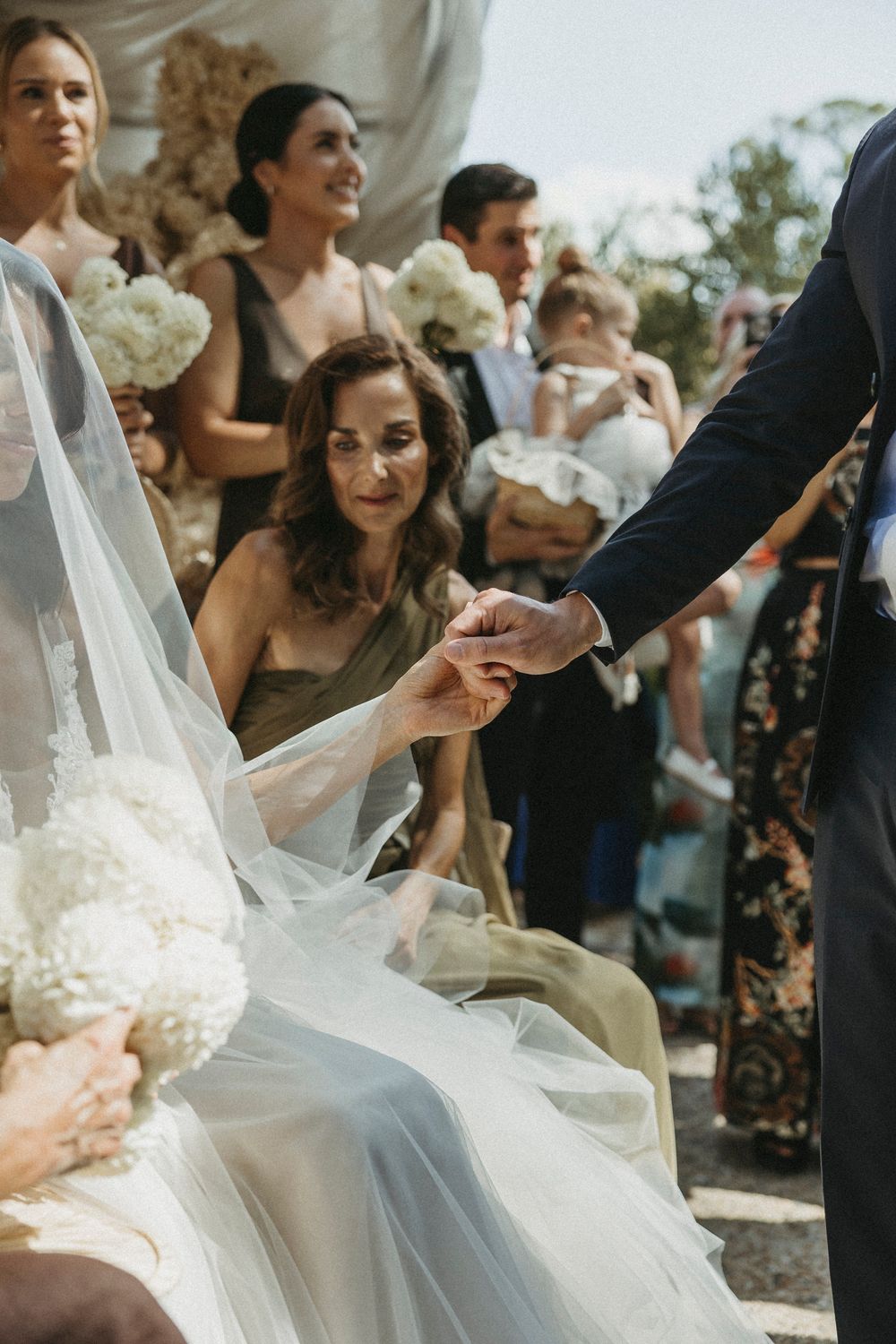 Bride in white veil holds grooms hand during intimate outdoor wedding ceremony moment.