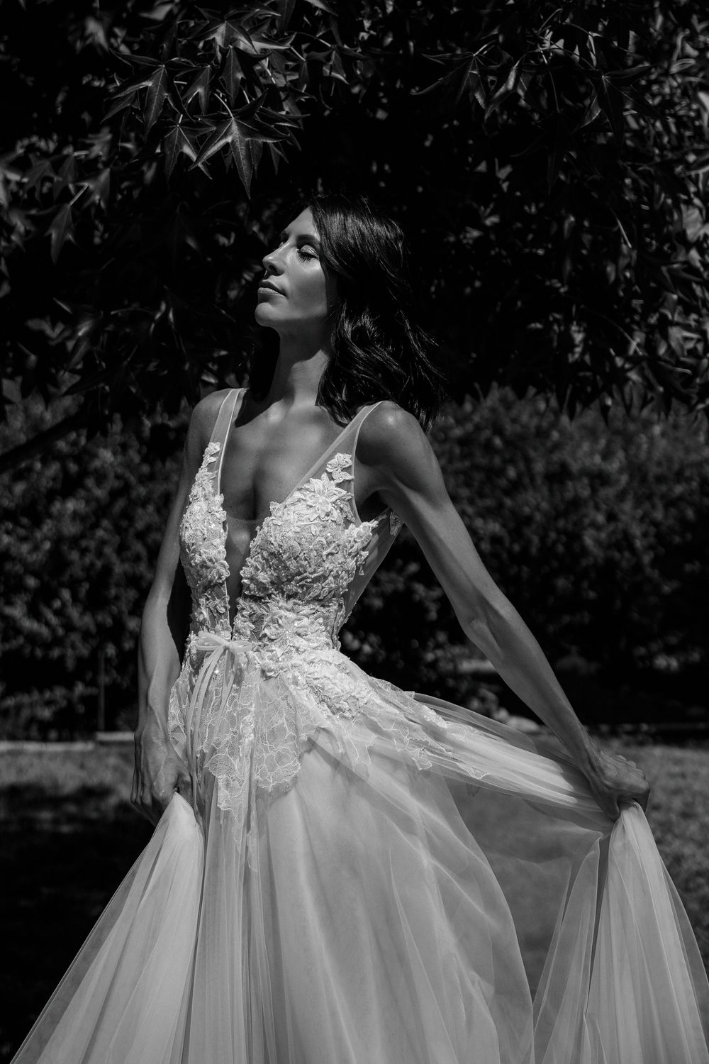 Black and white portrait of bride in floral lace gown posing under leafy tree branches.