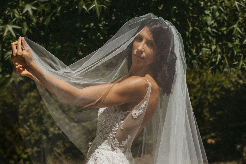 Bride in lace gown lifts sheer veil in sunlit garden setting.