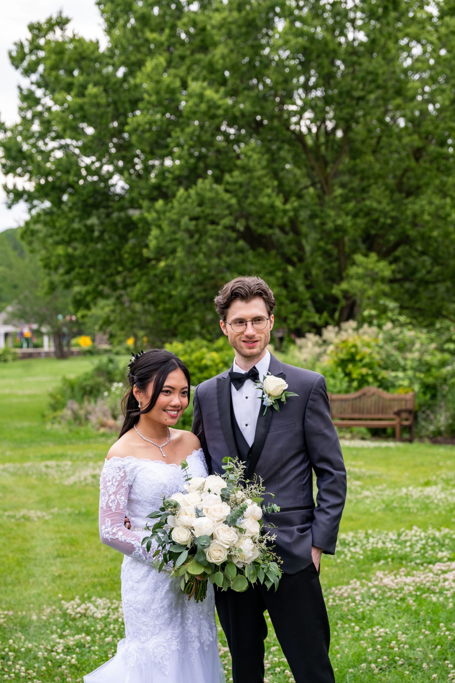 Bride and groom pose outdoors in lush garden, she holds white rose bouquet in lace gown, he wears gray tuxedo.