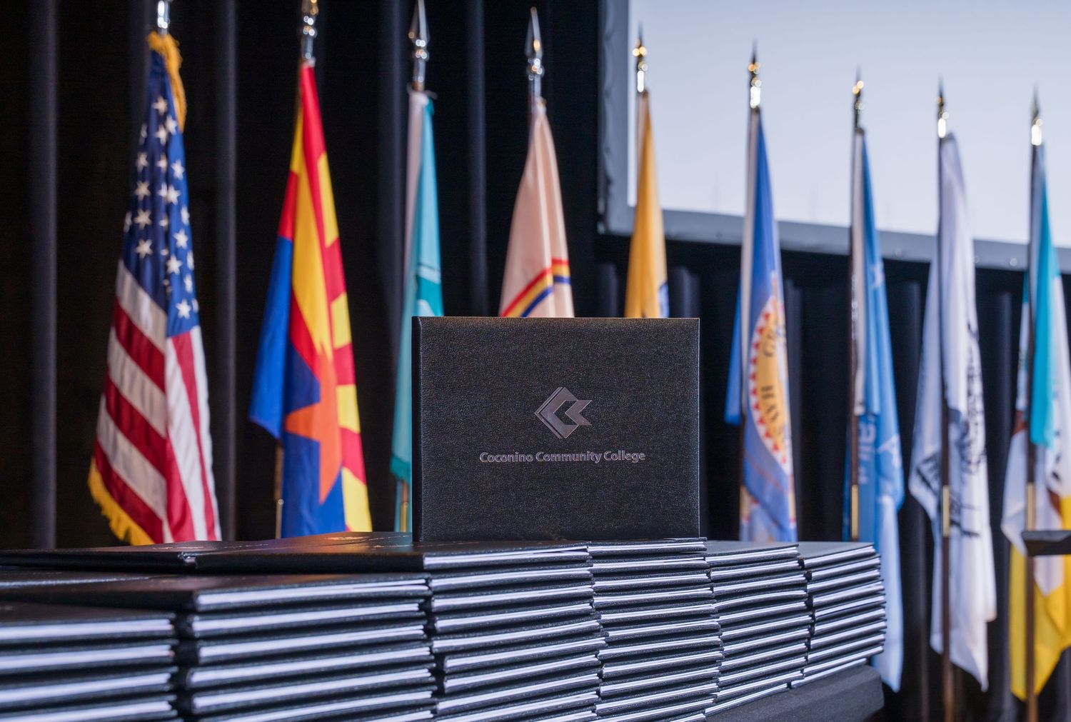 Chandler-Gilbert Community College diplomas stacked near a podium with colorful flags displayed on stage during graduation.