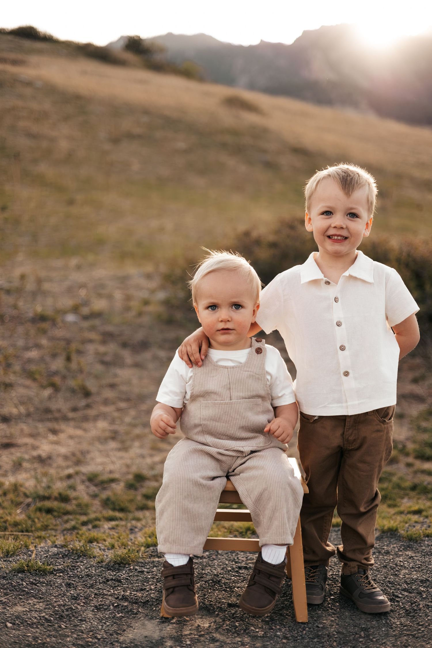 Two young brothers pose outdoors at golden hour, baby in overalls on a chair, older boy smiling in white shirt.