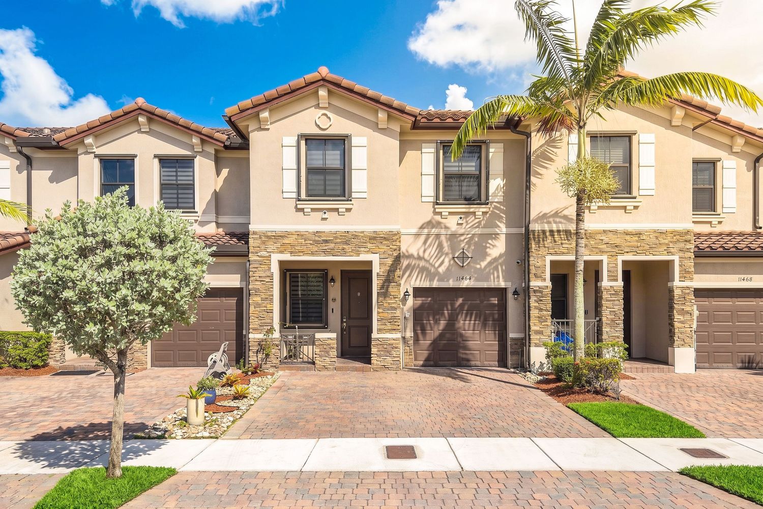 Two-story Mediterranean-style townhome with stone accents, brown garage doors, and brick paver driveway surrounded by tropical landscaping.