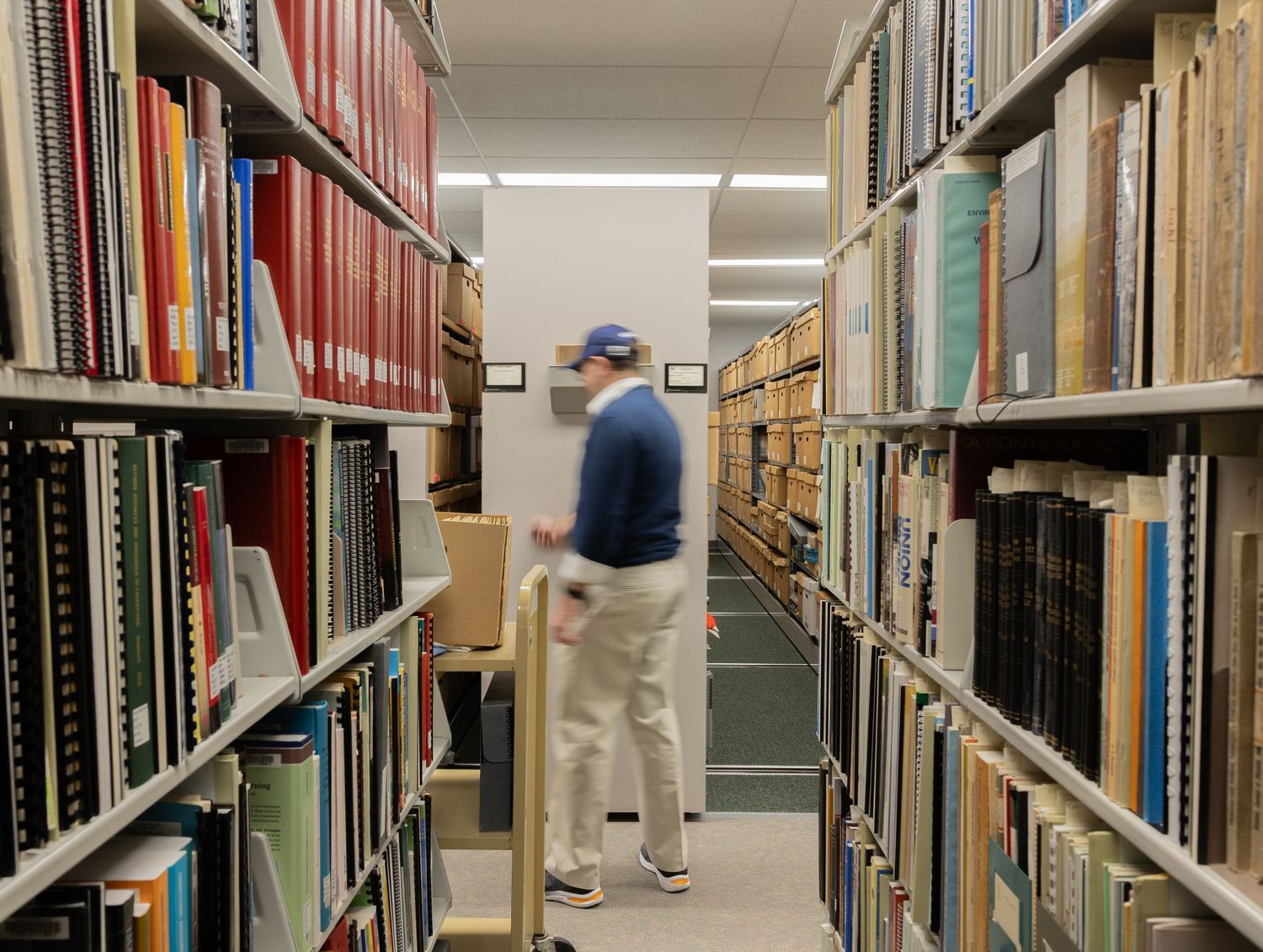 Person browsing through tall shelves filled with books and archival materials in a library stacks room.