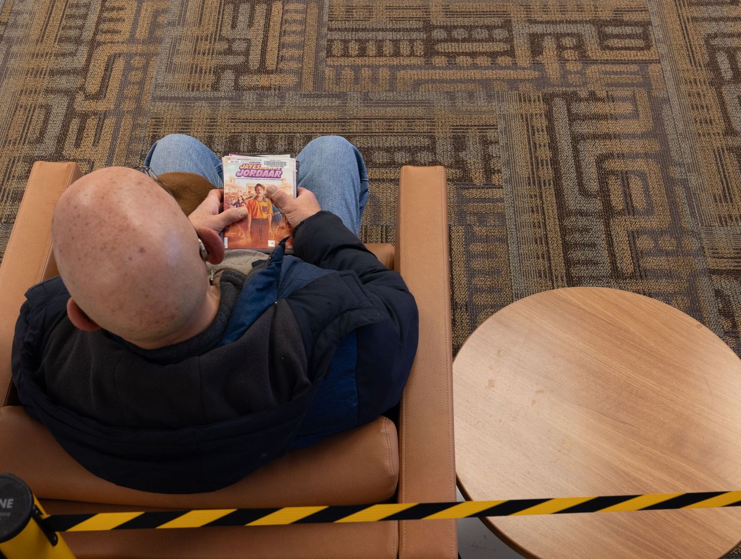 Overhead view of a bald man relaxing in a chair reading a magazine on patterned library carpet.