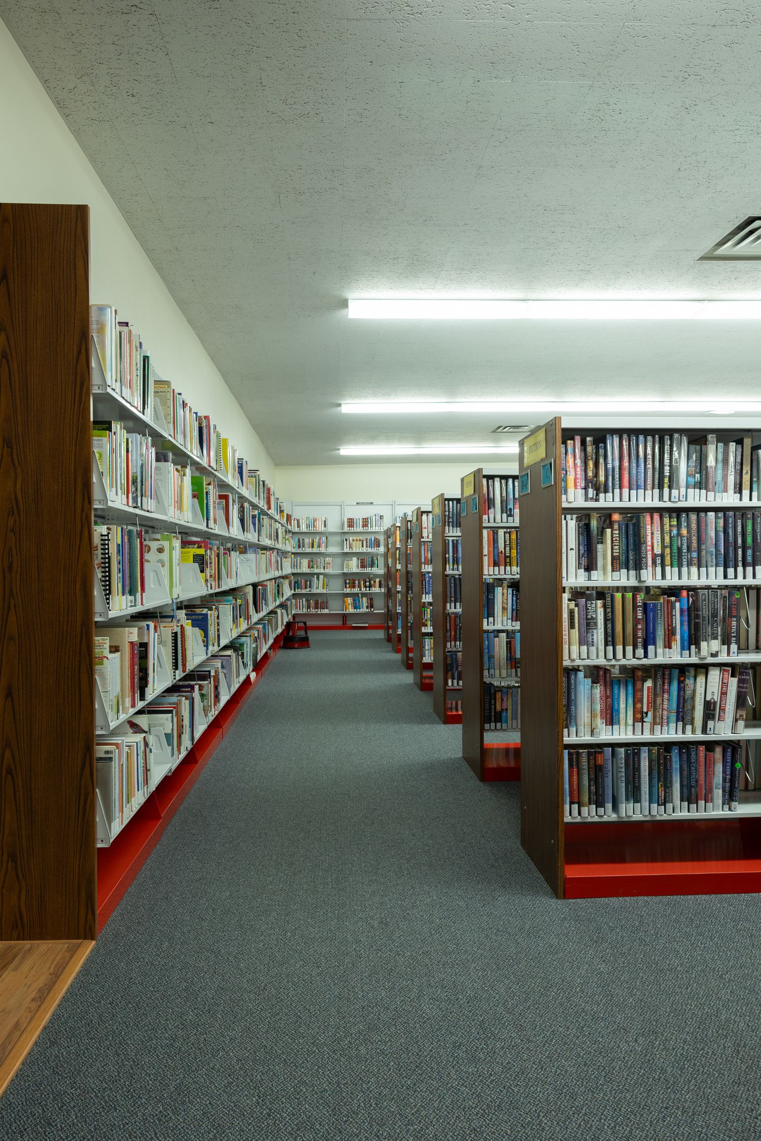 Long aisle between tall bookshelves filled with books in a well-lit public library with gray carpet.