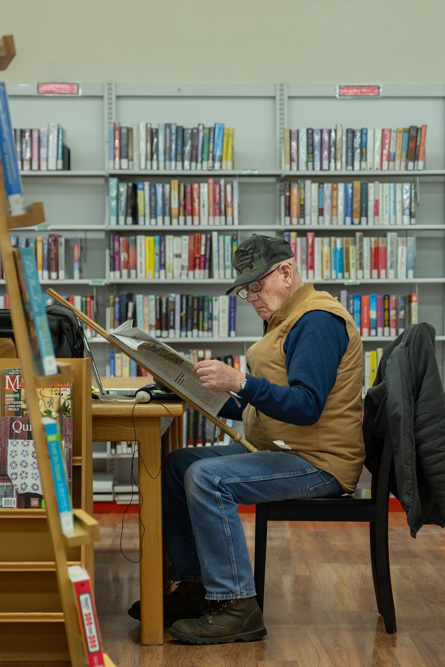 Man in cap and vest reading large book at library table surrounded by shelves of colorful books.