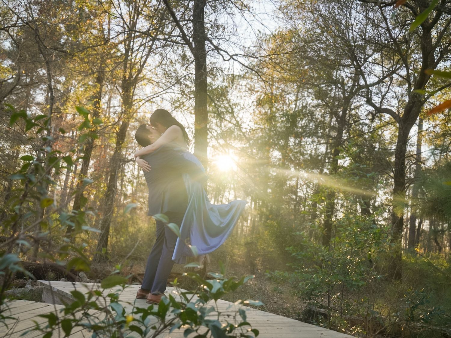 Couple embracing in sunlit forest clearing, golden rays streaming through autumn trees during romantic outdoor photoshoot.