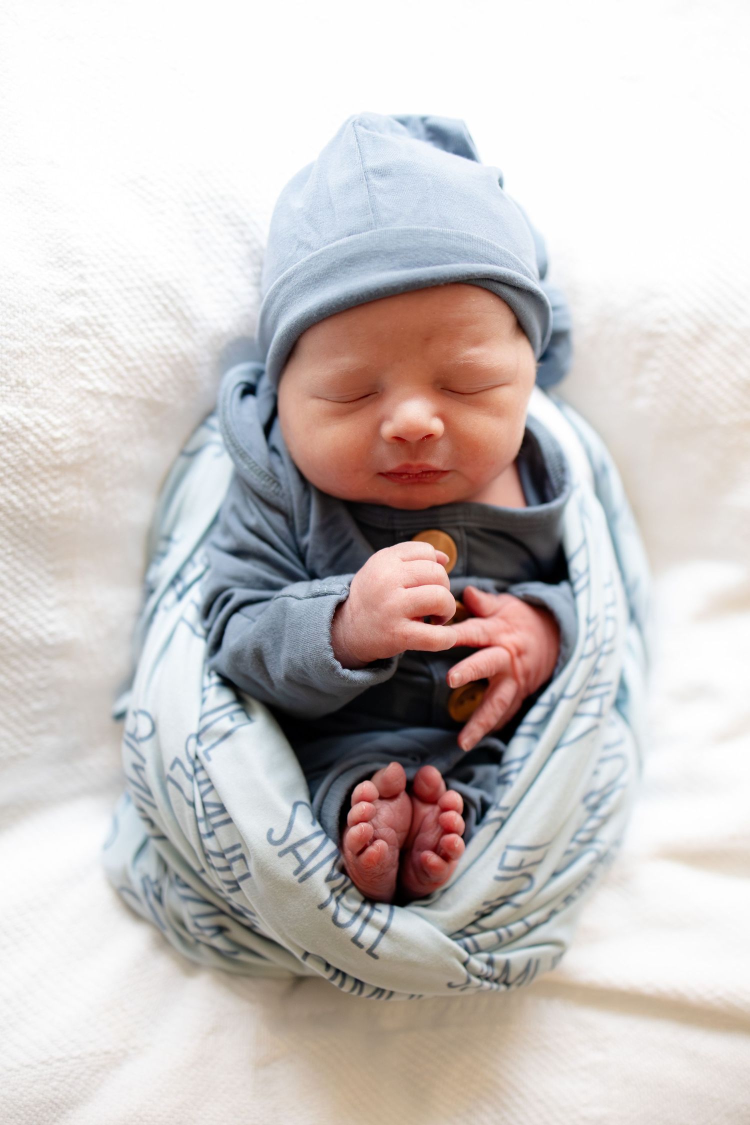 Peaceful newborn baby in blue sleeper and knotted hat rests on a white blanket.