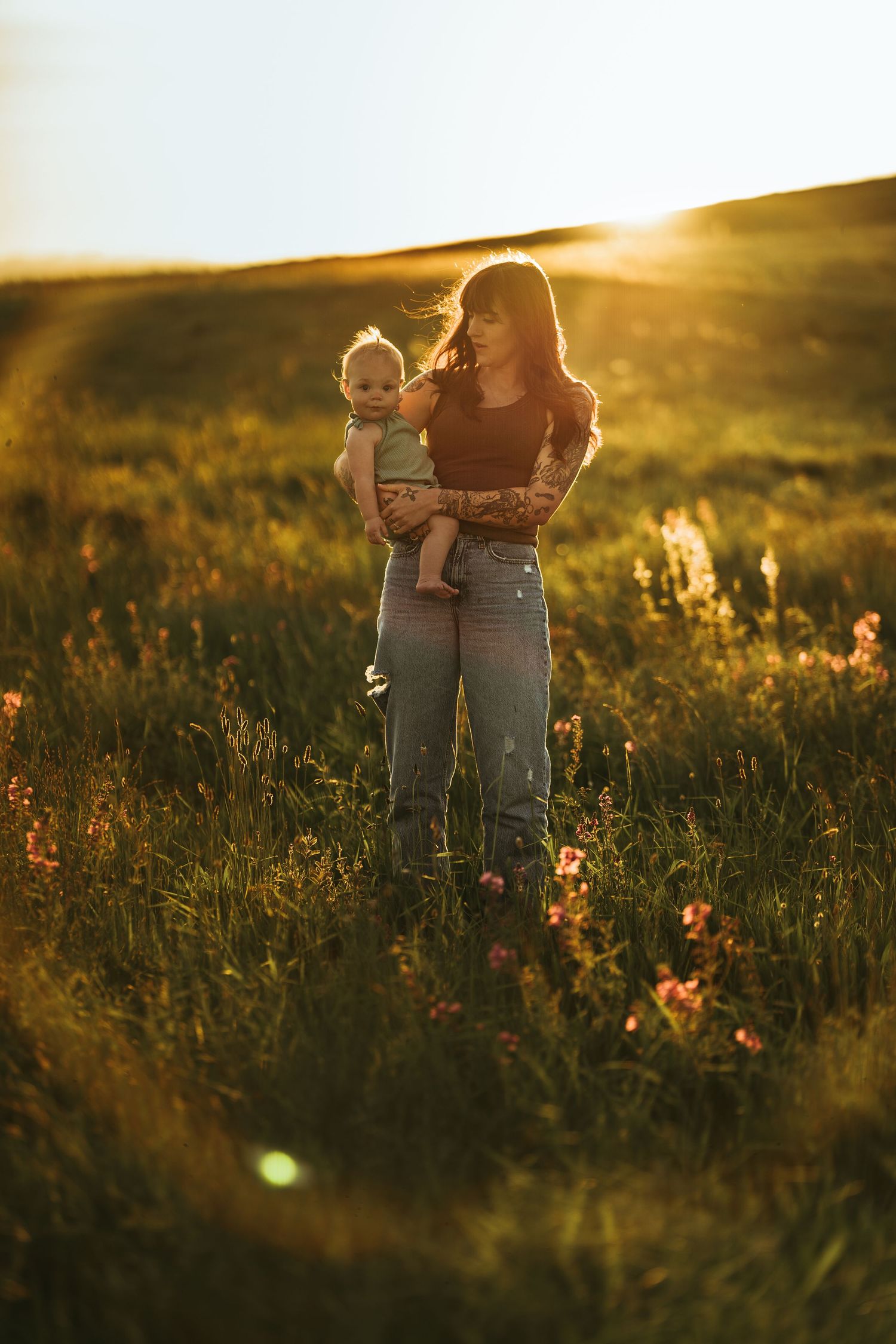 Tattooed mother tenderly holding her baby outdoors at golden hour in a green field.