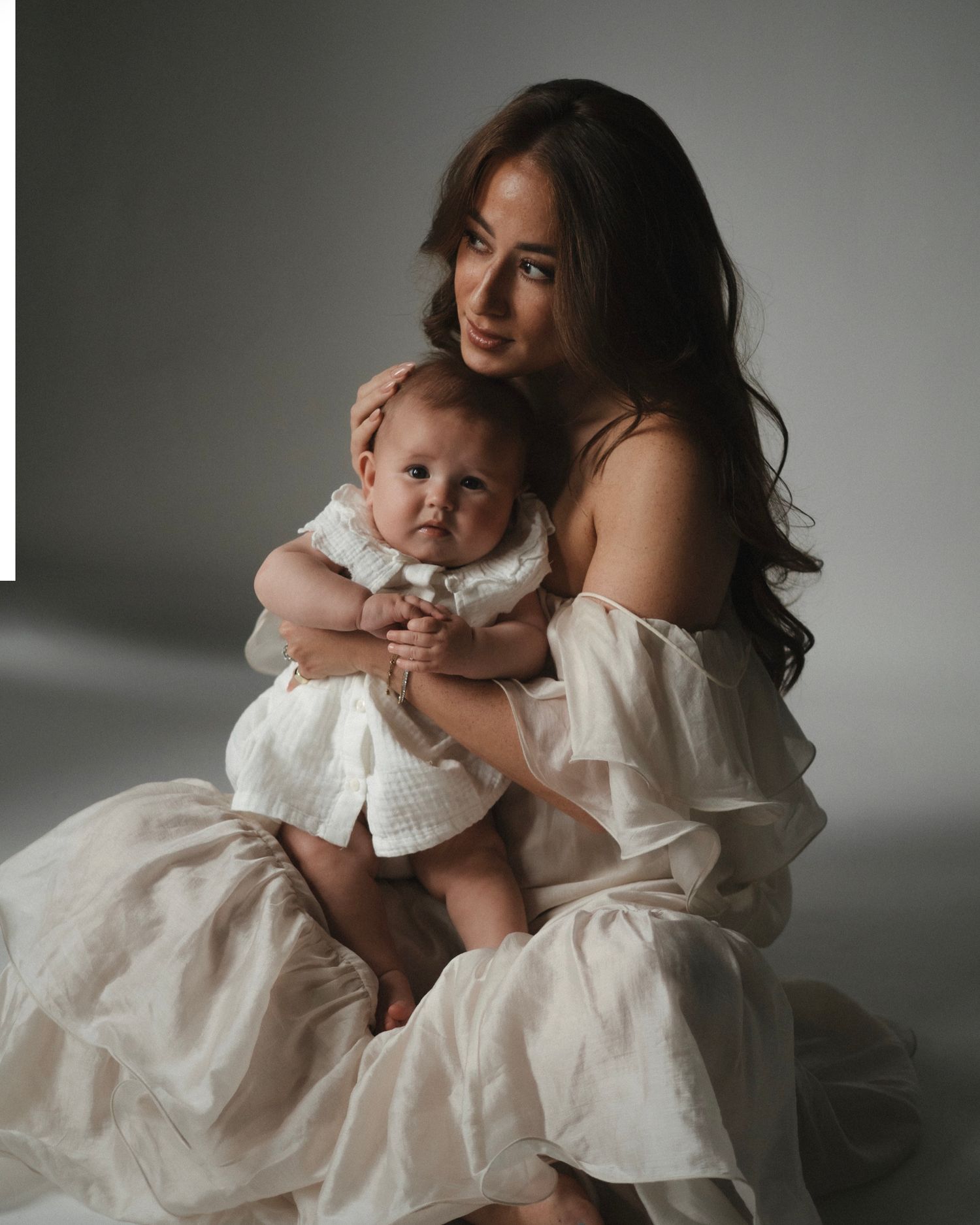 A woman in a white off-shoulder gown holds a baby in a white dress during an elegant studio portrait session.