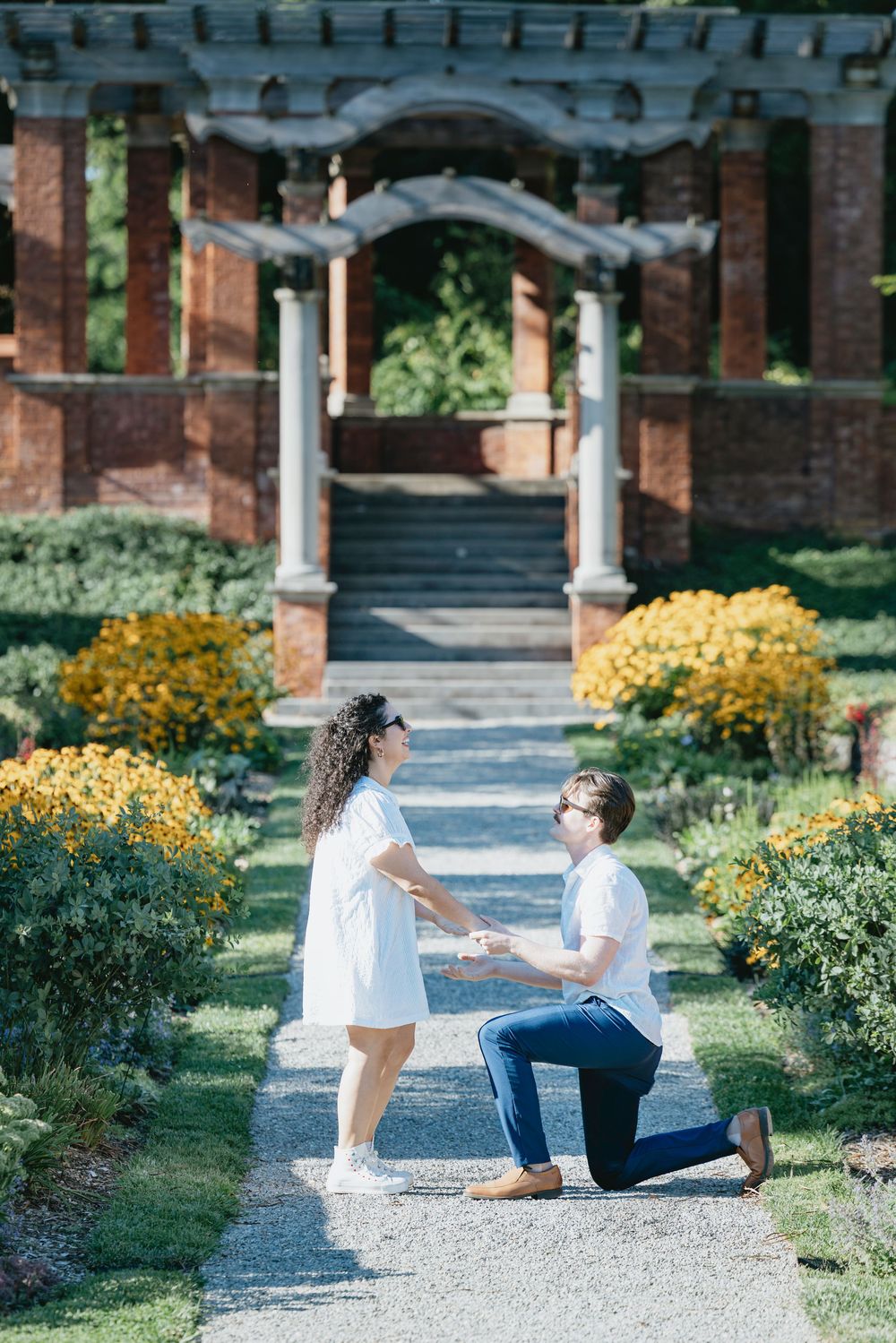 Man proposing on one knee to woman in white dress in a formal garden with yellow flowers and brick pergola.