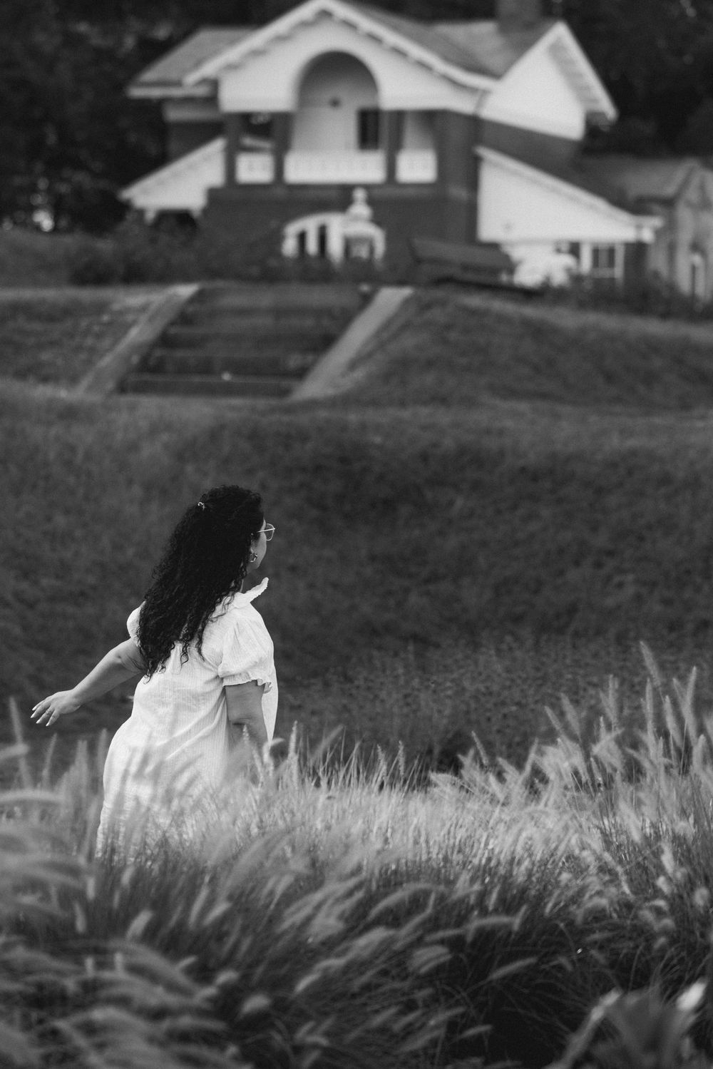 Black and white photo of a woman in a white dress walking through a field toward a house.