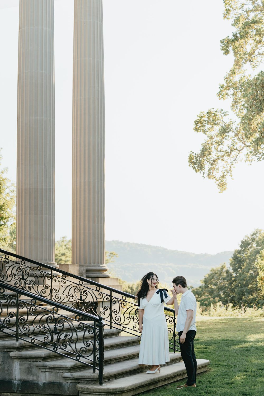 Couple standing on ornate stone steps beside tall columns in a lush green garden setting.