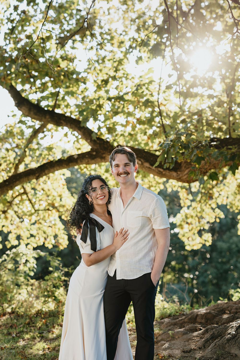 Couple smiling under sunlit tree, woman in white dress with black bow, man in cream shirt and black pants.