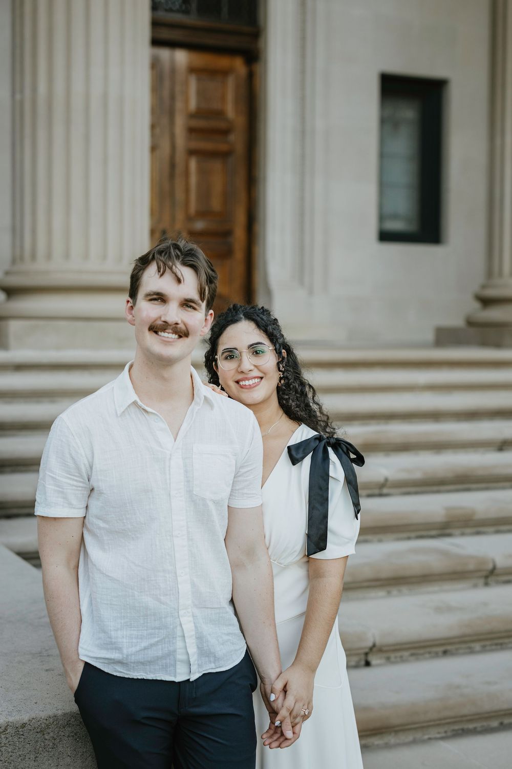 A smiling couple in white outfits holds hands on steps of a classical building with columns.