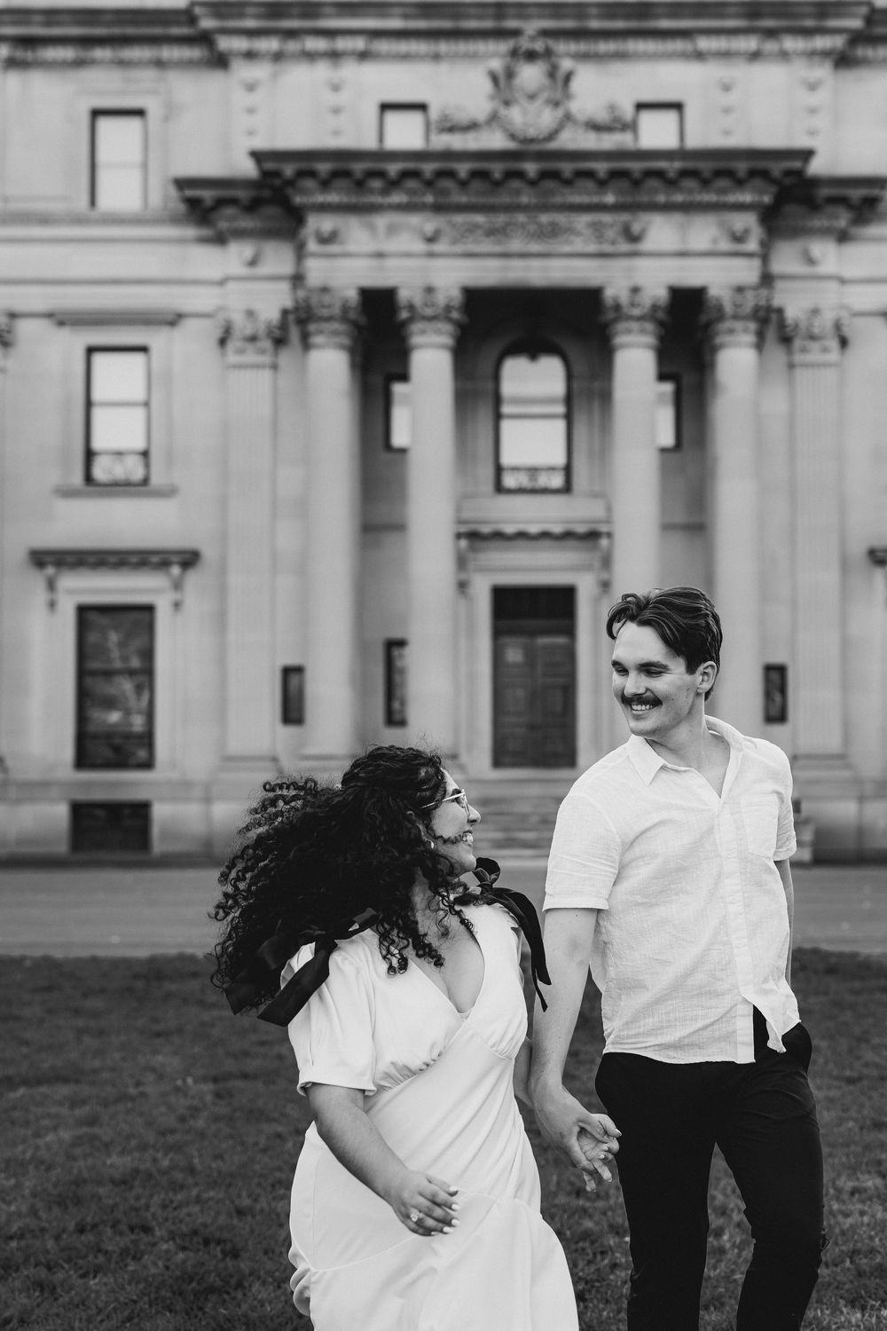 Black and white photo of a couple holding hands and smiling in front of a grand neoclassical building.