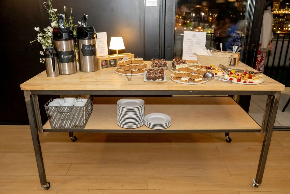 Buffet table on wheels with pastries, desserts, stacked plates, and stainless steel beverage dispensers at a catered event.