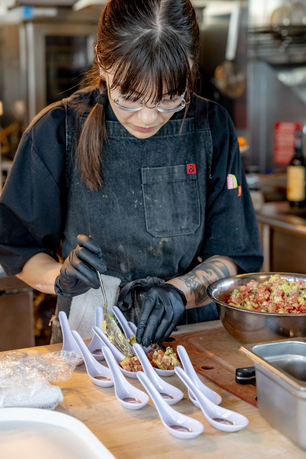 Female chef in black apron and gloves carefully plating appetizers on white spoons in a professional kitchen.