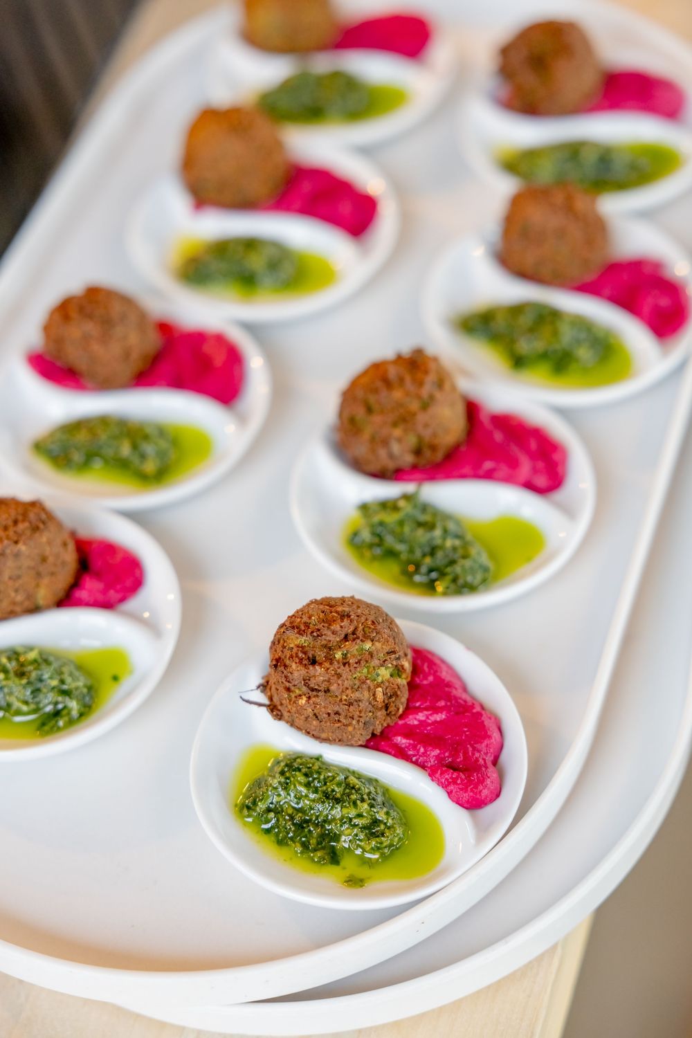 Elegant appetizer tray with falafel balls, vibrant beet hummus, and herb pesto sauce served on white spoons.