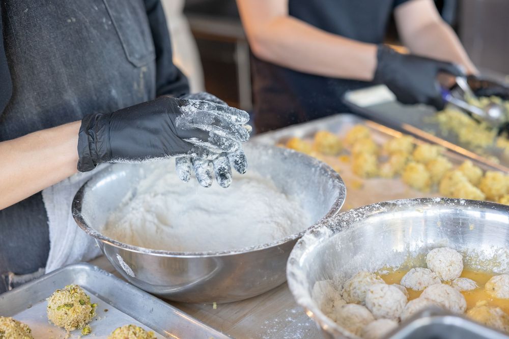 Chef in black gloves kneading white dough in a steel bowl surrounded by breaded food balls in a commercial kitchen.
