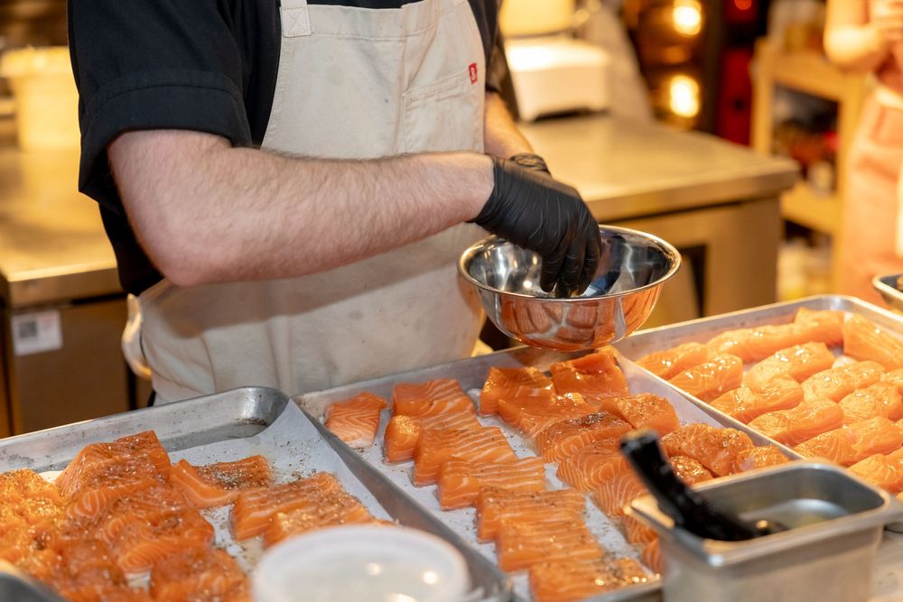 Chef in apron and black gloves preparing fresh salmon fillets on a metal tray in a seafood market.