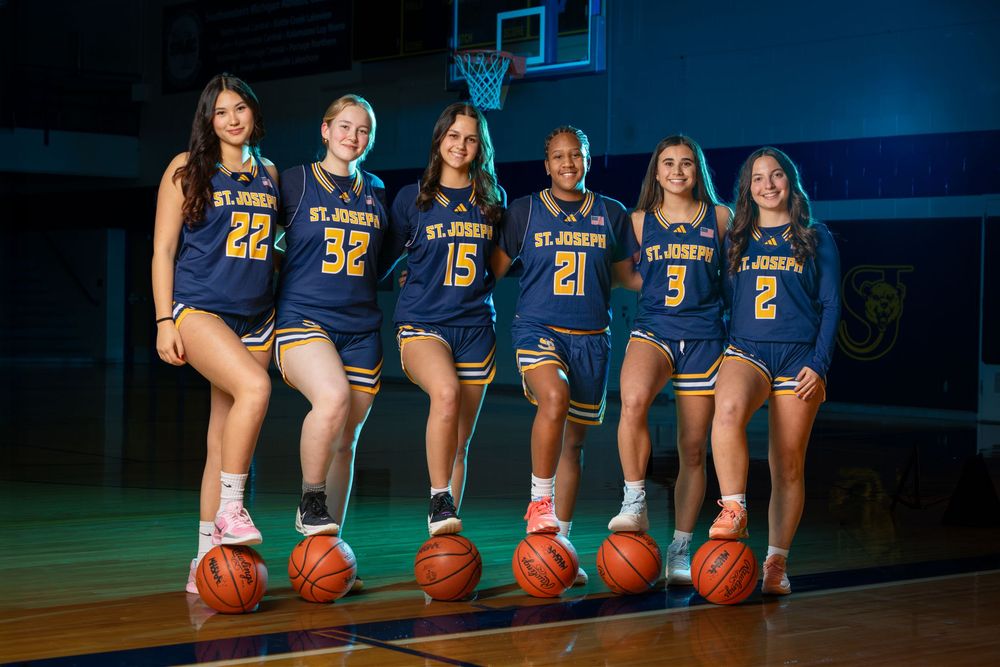 Six St. Joseph girls basketball players in navy uniforms pose with basketballs on a gym court.