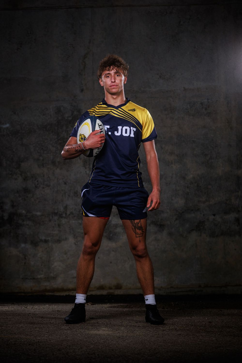 Young male rugby player in navy and yellow St. Joe jersey holds a rugby ball, posing against a dark concrete wall.