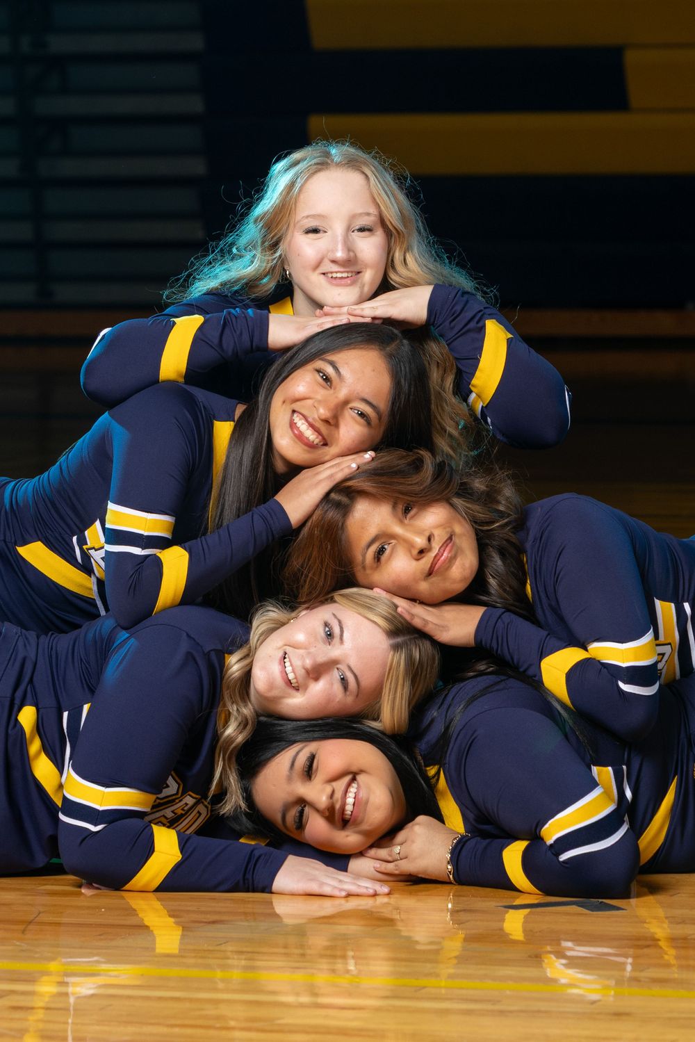 Five cheerleaders in navy and gold uniforms pose in a pyramid stack on a gym floor, smiling at the camera.
