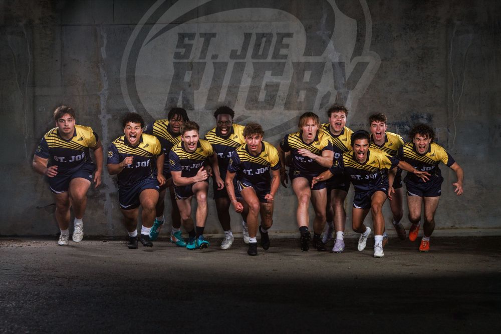 St. Joe Rugby team poses dynamically in matching blue and gold uniforms against a concrete wall with team logo.