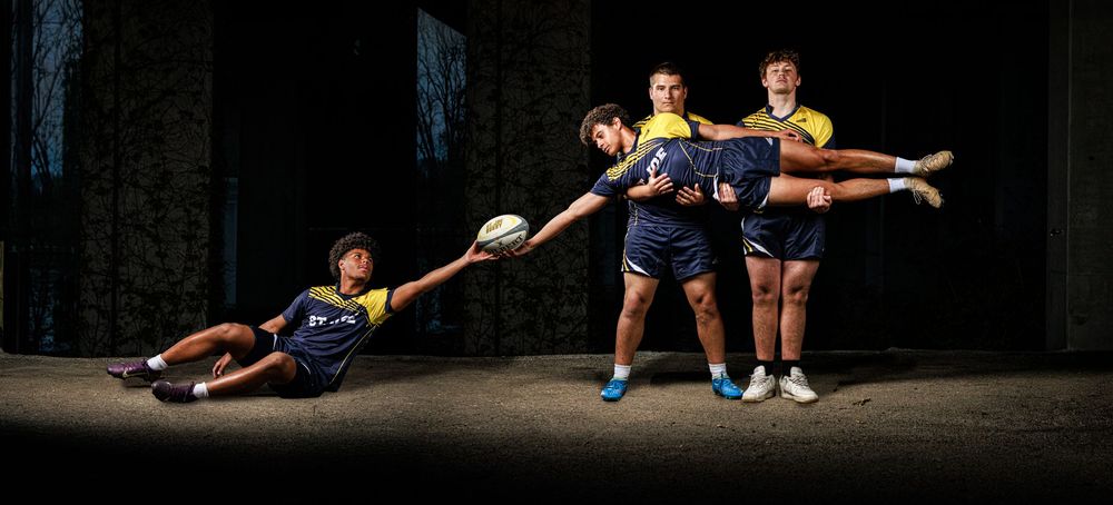 Four rugby players in blue and yellow uniforms perform a dynamic action pose passing a ball against a dark background.