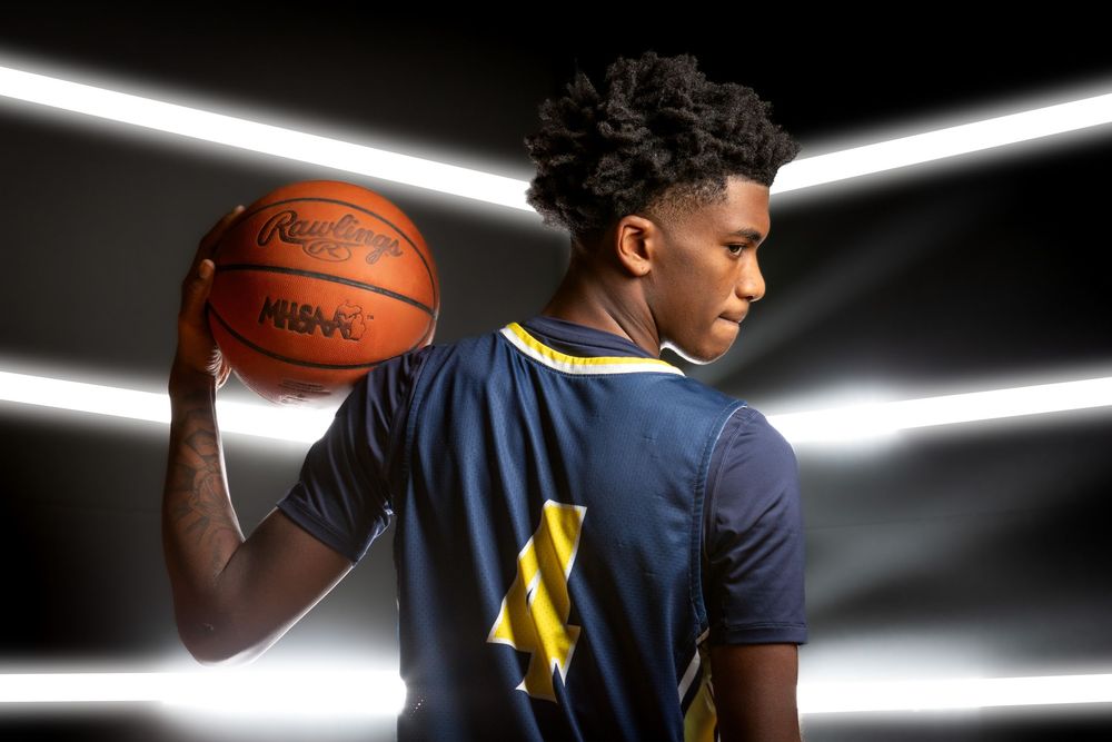 Young male athlete holding a basketball on his shoulder, wearing a navy and gold jersey, posed against a dramatic lit background.