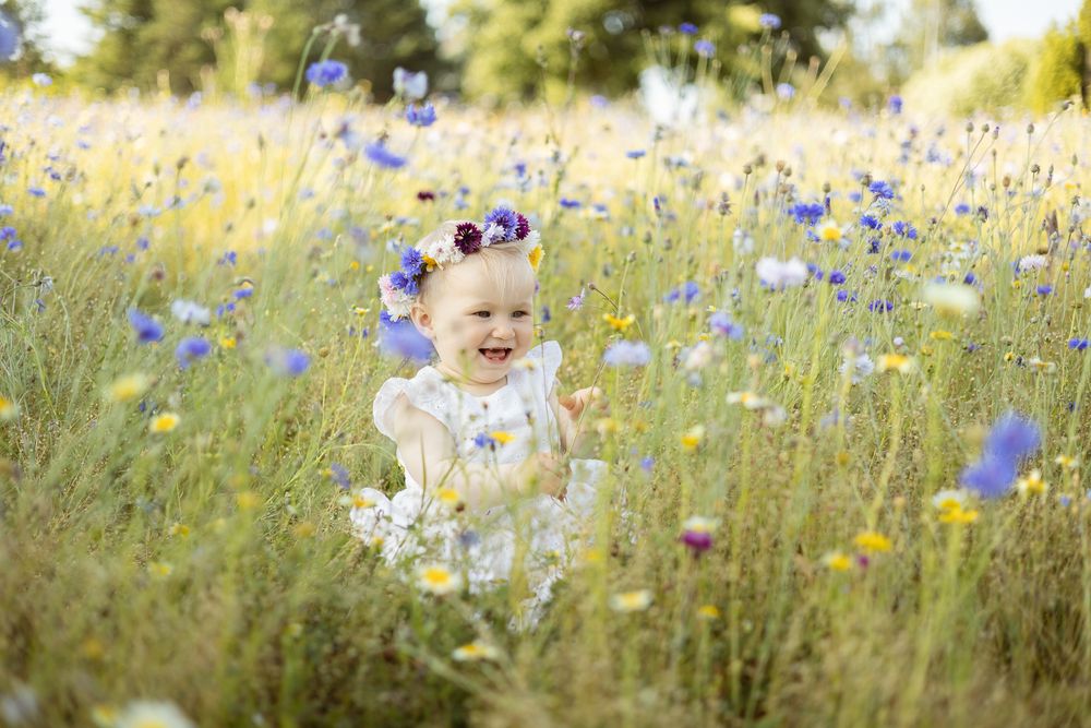 alt="A joyful baby girl in a white dress and floral crown laughing in a sunny wildflower meadow"