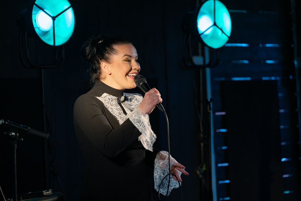alt="Woman in black dress with lace detail smiles while holding microphone on stage with teal lights"